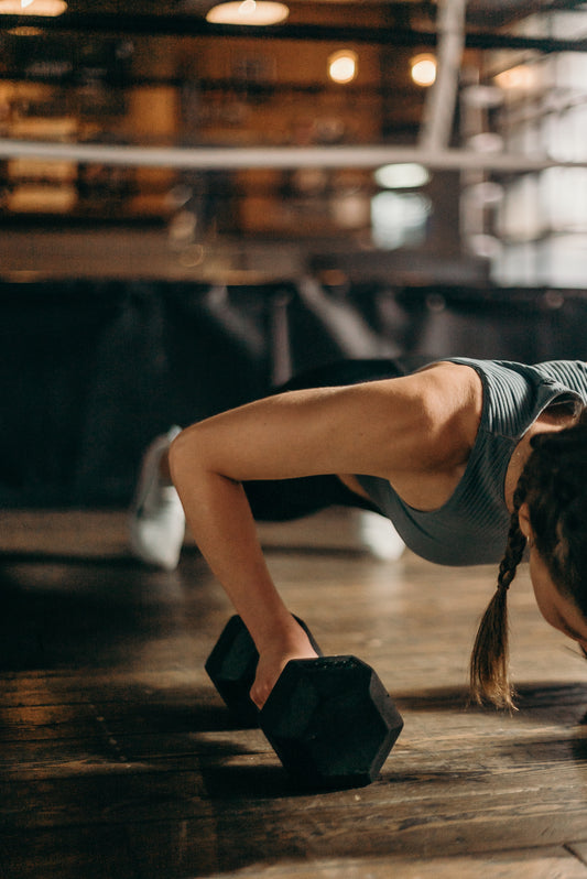 In the plank position, a woman performs dumbbell exercises on a wood floor